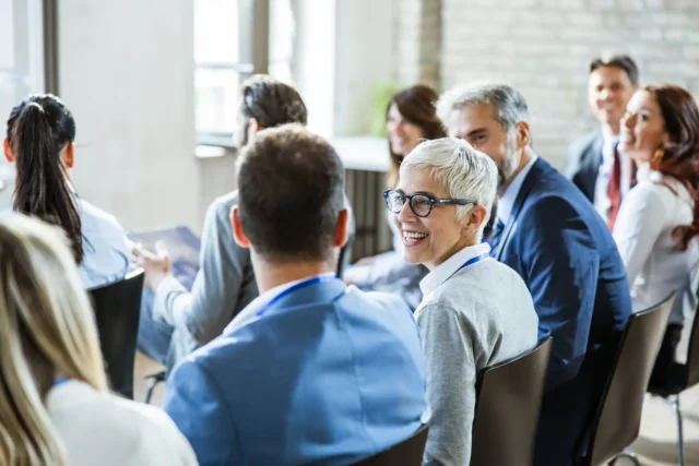 A diverse group of professionals, including a cheerful senior woman with short white hair and blue glasses, engaged in a Medicare information session. They are seated, facing the front of a well-lit conference room, indicating an atmosphere of active learning and collaboration relevant to a Medicare agency