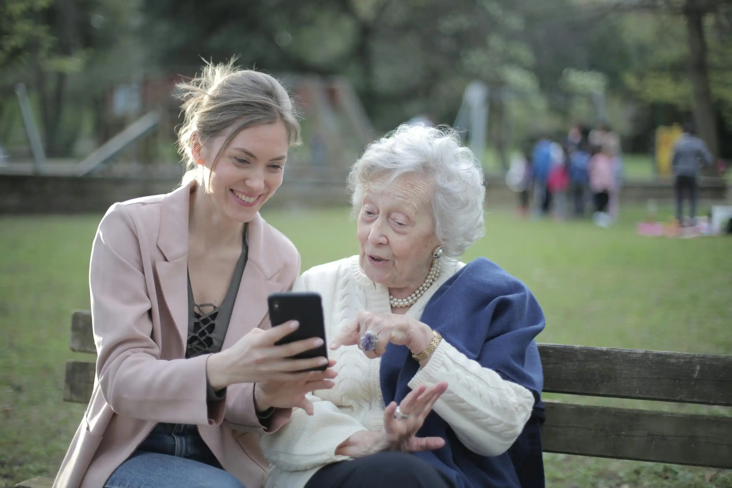 A young woman and an senior woman are sitting together on a park bench, smiling as they look at Medicare plans on a smartphone screen held by the younger woman