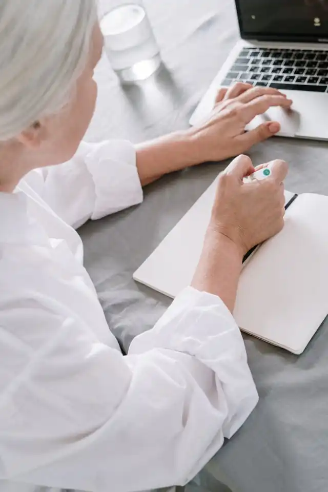 Over-the-shoulder view of a senior woman wearing a white blouse using a laptop on a grey tablecloth, with a notepad and pen at hand and a glass of water nearby, possibly researching or gathering information about Medicare plans